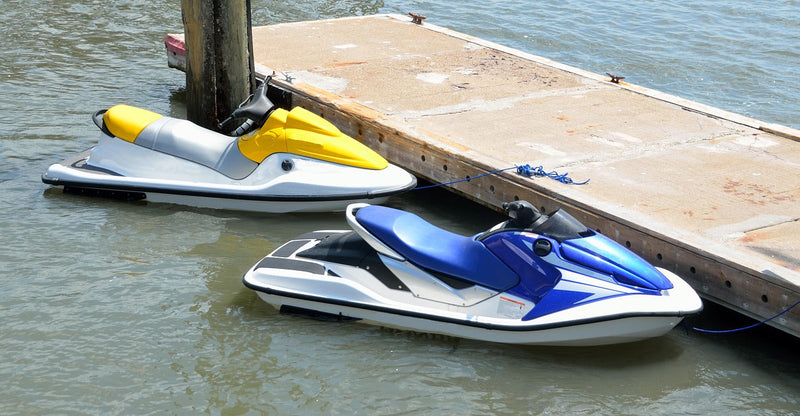 Two jet skis (yellow and blue) tied with ropes to a traditional fixed wooden pier, contrasting with the convenience of modern floating docks for jet ski